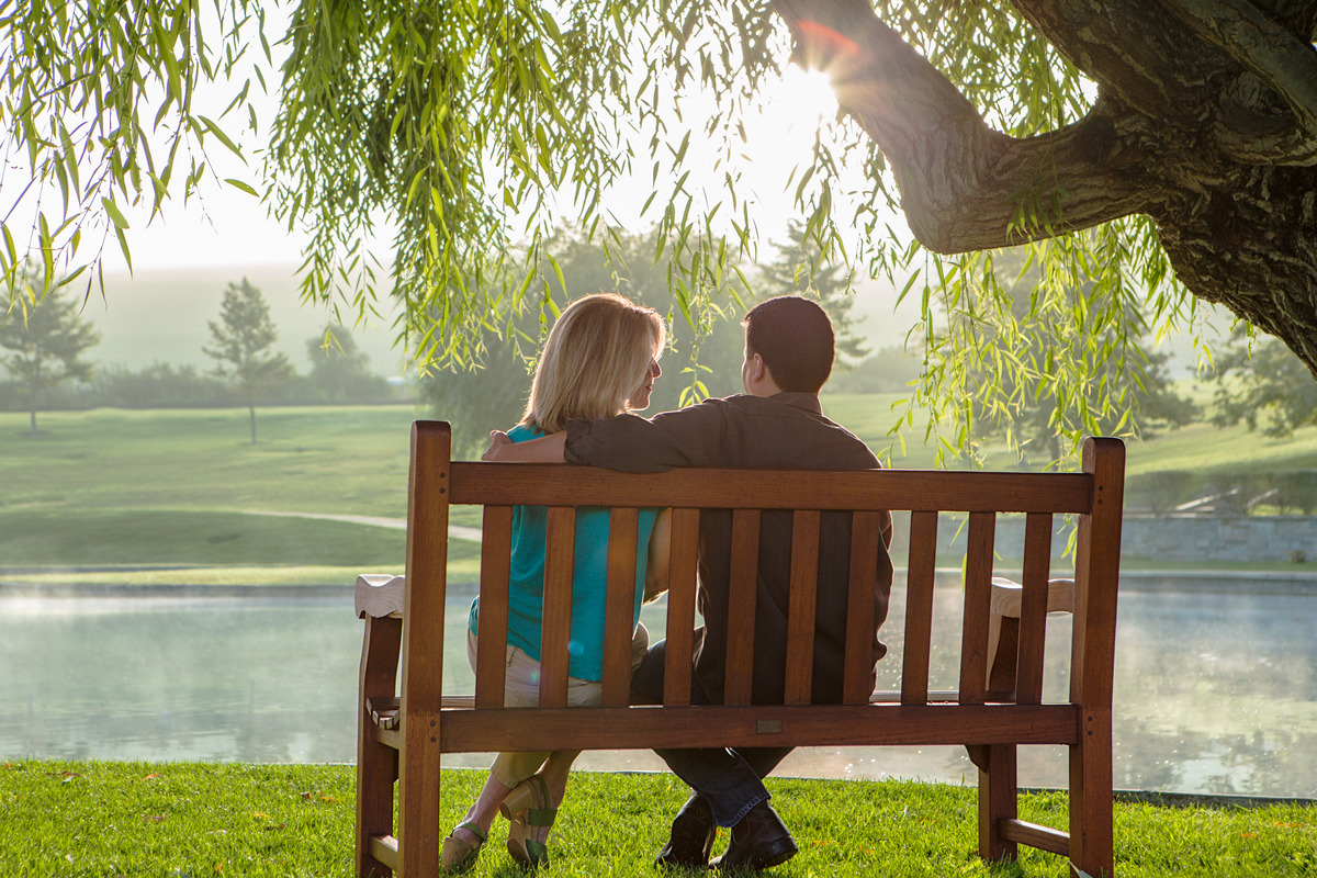 Couple sitting on a wooden bench in front of a lake underneath a tree.