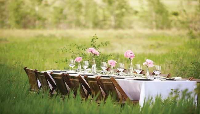 A table with a white table cloth set with glasses and plates in a green meadow