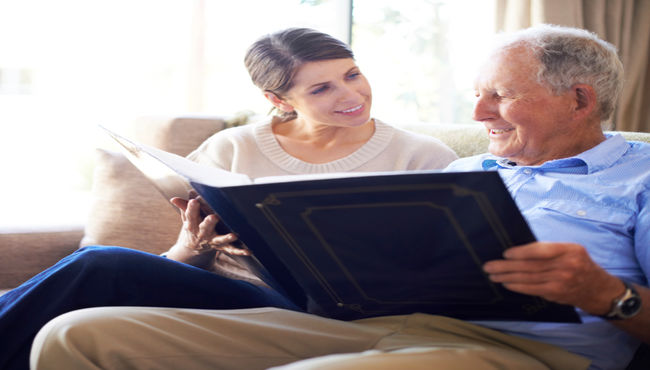 A senior man in a blue shirt smiles as he reminisces with his adult daughter while looking at a large photo album. 