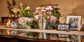 Mesa con una urna de metal, cuadros, flores y una vela encendida en Pacific View.