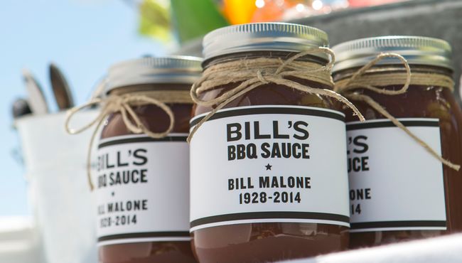 Jars of Bill's BBQ sauce sit on a tray at an outdoor BBQ funeral celebration.