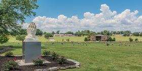 Statue of praying hands on cemetery grounds with a mausoleum in the background at Sherwood Chapel and Memorial Gardens. 