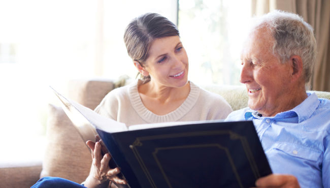 A senior man in a blue shirt smiles as he reminisces with his adult daughter while looking at a large photo album. 