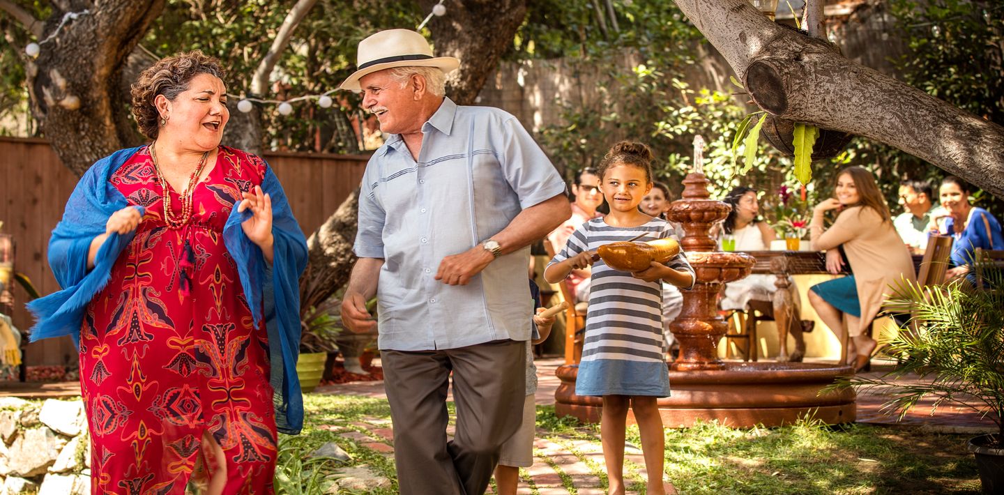 A cheerful senior couple dances together outside in a sunny courtyard as family looks on.