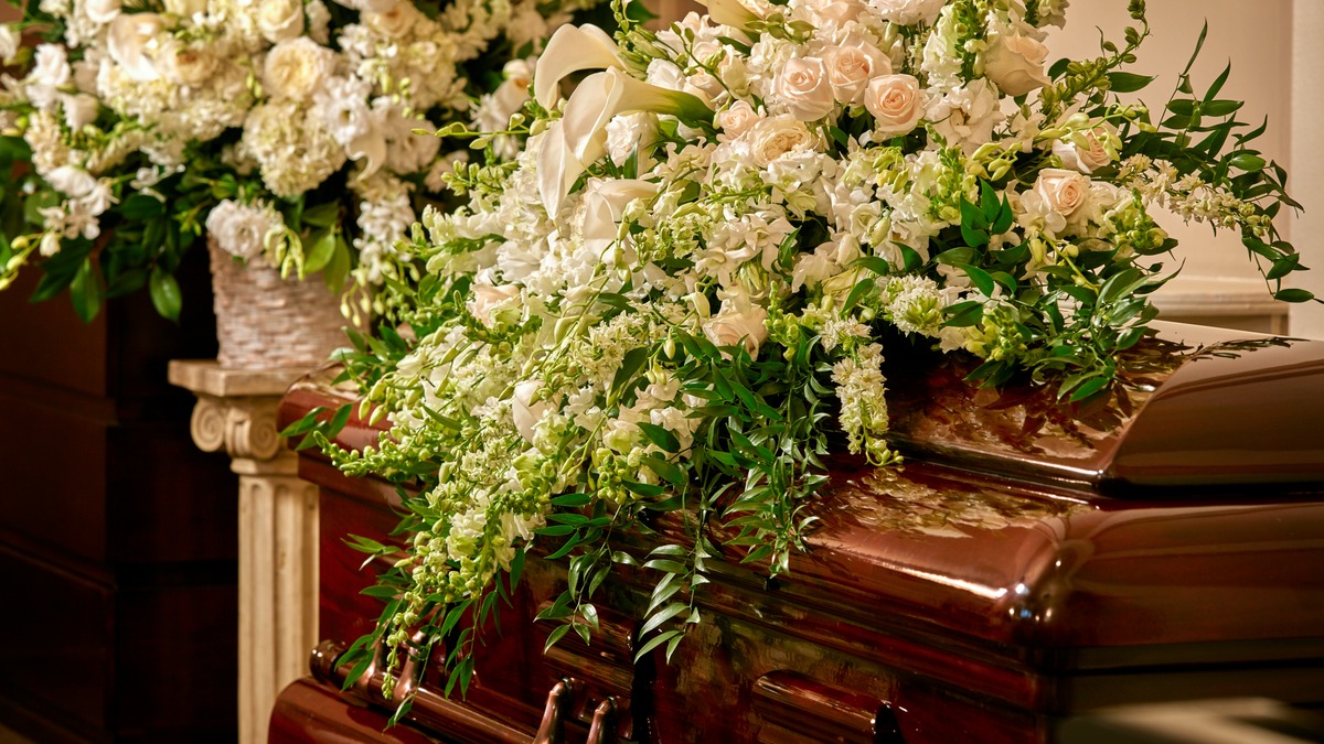 Spray of white roses and tulips laid on top of a wooden casket inside a funeral home. 