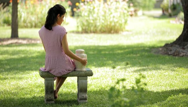 A young woman sits and reflects on a garden bench with a marble urn