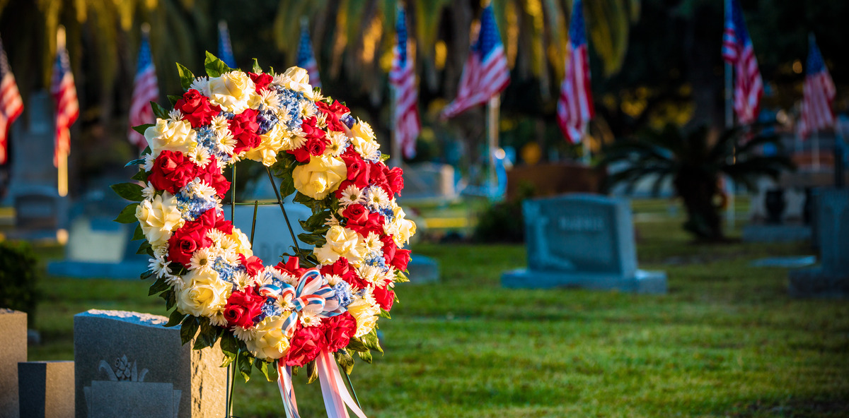 A patriotic floral wreath is displayed in a cemetery with upright markers and American flags