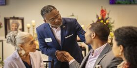 A funeral director shakes the hand of a seated man at a reception